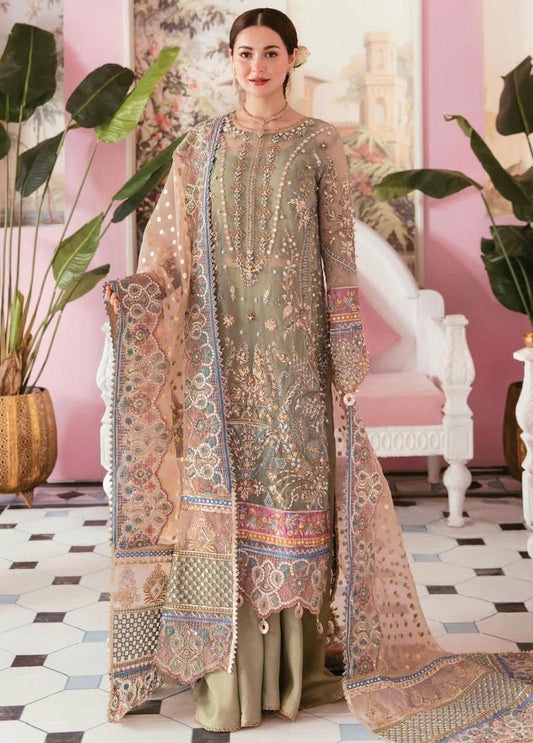 Woman wearing an olive green embroidered shalwar kameez with organza dupatta, showcasing moti, sheesha, and latkan detailing, standing in a richly decorated indoor setting.