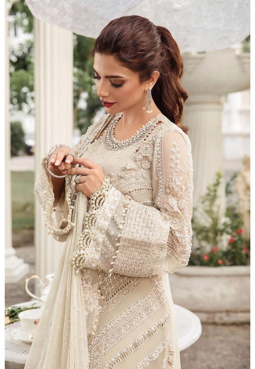 Woman wearing a beige Maria B chiffon suit with full embroidery, cutwork, and tassel detailing. Outfit includes chiffon dupatta and malai trouser, styled in an elegant garden setting.